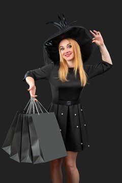 Young woman smiling in a black witch hat and dress, confidently holding multiple dark shopping bags, presenting a concept of holiday shopping, Halloween sales, or seasonal promotions