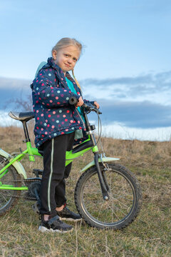 Blonde little girl with pigtails carefully holding handlebars of her green bicycle, looking at the camera while standing on dry grass under a blue sky