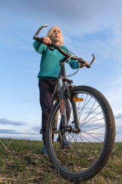 Young blonde person standing with a bicycle on a grassy hill, holding the handlebars with a determined expression, looking forward towards a blue sky representing freedom and future aspirations