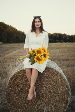 woman in a white dress holding a bouquet of wild flowers with sunflowers in a mowed field with straw bales