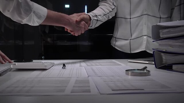 Business professionals shake hands over a cluttered desk of contracts, calculator and magnifying glass, sealing a trusted partnership after detailed financial review and late night work