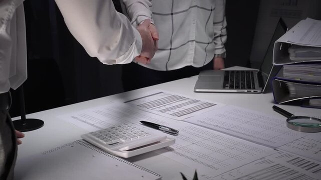 Business professionals shake hands over a cluttered desk of contracts, calculator and magnifying glass, sealing a trusted partnership after detailed financial review and late night work