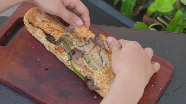 hands opening sliced fresh submarine sandwich to reveal melted cheese and meat filling on wooden board, outdoor background, horizontal.