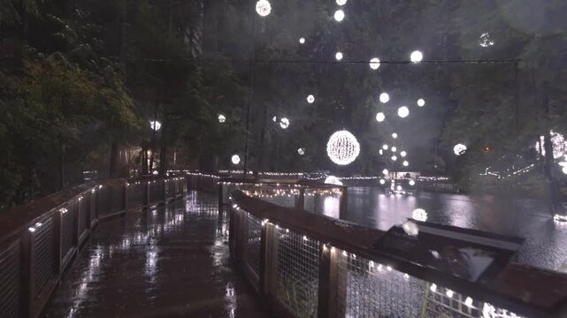Enchanting rainy night path with glowing festive lights suspended over wet wooden boardwalk at Capilano suspension bridge park.