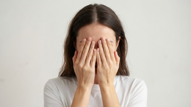 Sad brunette woman covering her face with hands experiencing deep emotional pain while standing against a neutral plain white background