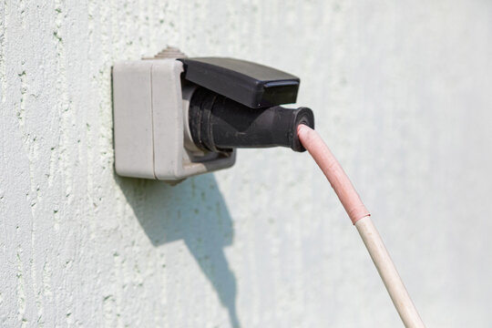 Waterproof electrical socket mounted on a white exterior wall with an inserted power cable. Industrial equipment for outdoor electricity supply on construction site and building