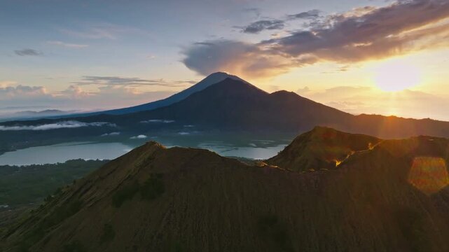 Aerial view showing the volcanic crater of mount Batur with lake Batur and mount Agung in background, illuminated by the golden light of sunrise over the Indonesian landscape. Drone flight panorama