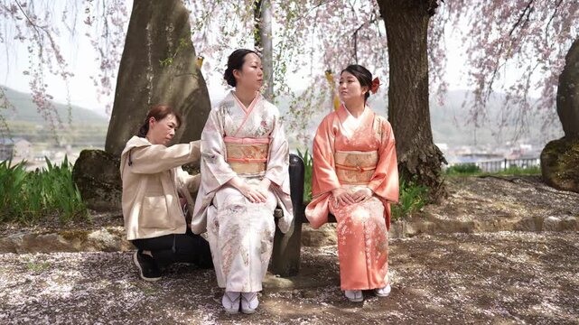 An assistant kneels beside two women on a bench and adjusts the back of a patterned kimono under cherry blossoms. Japan, April 19, 2025, spring.