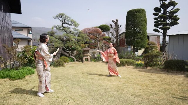 Two women in kimonos play hanetsuki with wooden paddles as a shuttlecock flies above a landscaped garden lawn. Japan, April 19, 2025, spring.