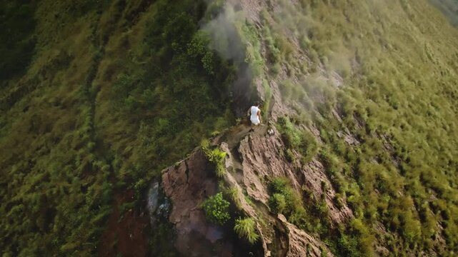 Man running on a narrow, rocky ridge of mount Batur, navigating the challenging terrain covered with green vegetation, embodying active sports, adventure and exploration in nature. Aerial drone shot