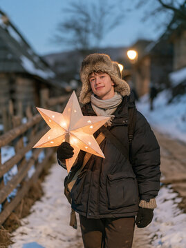 Young man in an ushanka hat holding a decorative light star in a winter village