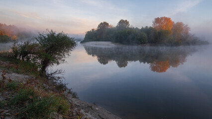 Obraz premium calm, misty river reflecting lush green and autumn-colored trees under a soft, hazy sky.