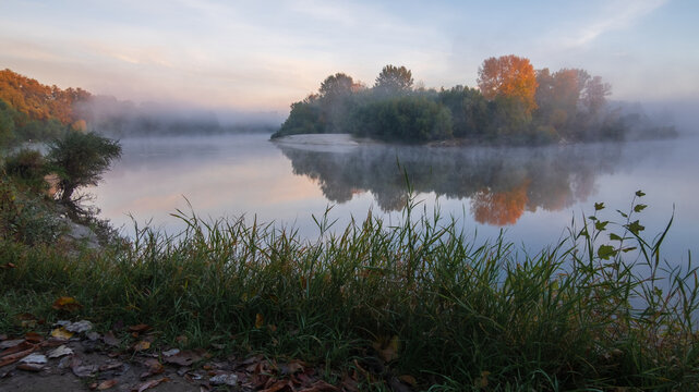 A serene, misty morning landscape shows a calm river reflecting autumn trees on a small island under a soft sky.