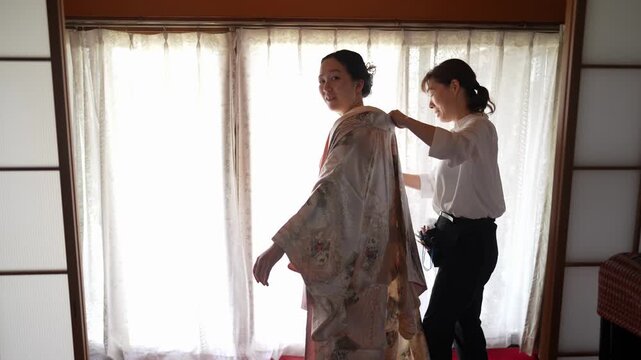 An assistant arranges a patterned kimono on a woman seen from behind beside a bright window. Japan, April 19, 2025, spring.
