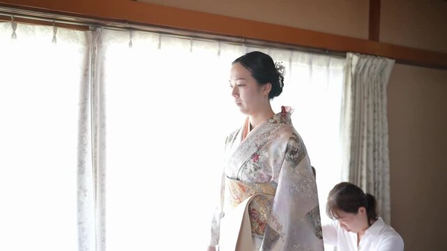 A woman stands in profile while an assistant works at the back of her patterned kimono. Japan, April 19, 2025, spring.