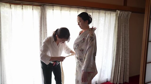 An assistant bends forward to secure the waist of a woman's patterned kimono beside a window. Japan, April 19, 2025, spring.