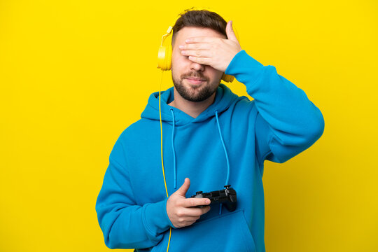 Young caucasian man playing with a video game controller isolated on yellow background covering eyes by hands. Do not want to see something