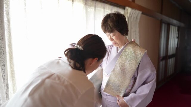 An assistant wraps a pale patterned obi around a woman wearing a lavender kimono. Japan, April 19, 2025, spring.