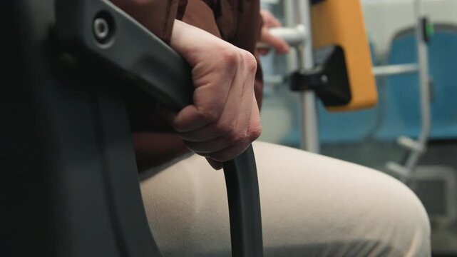 Close low angle view of seated adult holding armrest and vertical pole with both hands, showing steady posture, secure grip, and calm balance inside public transit cabin during urban travel