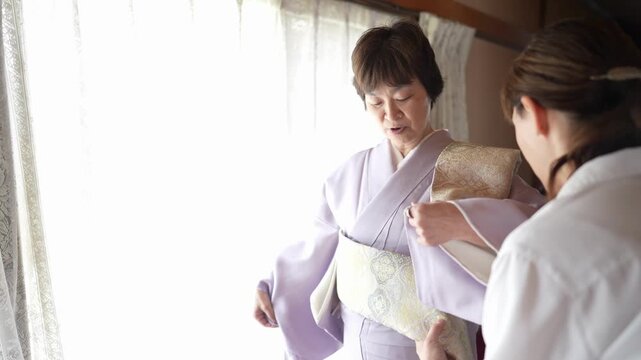 A woman in a lavender kimono looks toward the camera as an assistant adjusts her pale patterned obi. Japan, April 19, 2025, spring.