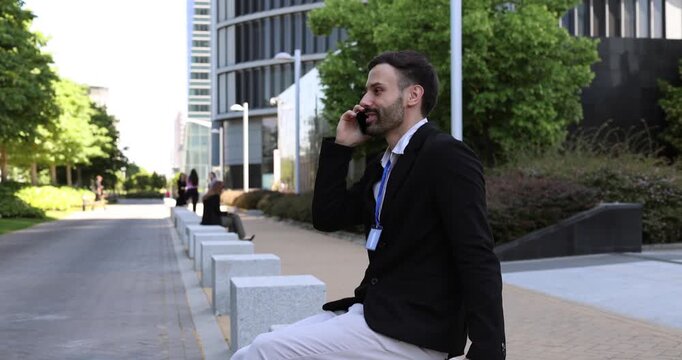 Young businessman wearing an id badge sitting outside in front of a modern building, smiling while talking and gesturing on his mobile phone during a sunny day
