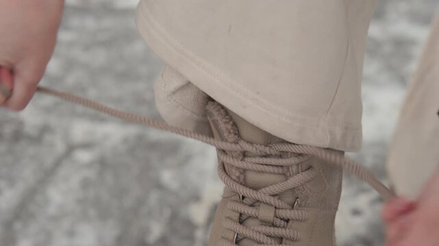 Hands actively lace, adjust, and secure tan combat boot with neat bow resting on gray concrete pavers, showing close detail of footwear texture, stitching, eyelets, and practical outdoor wear