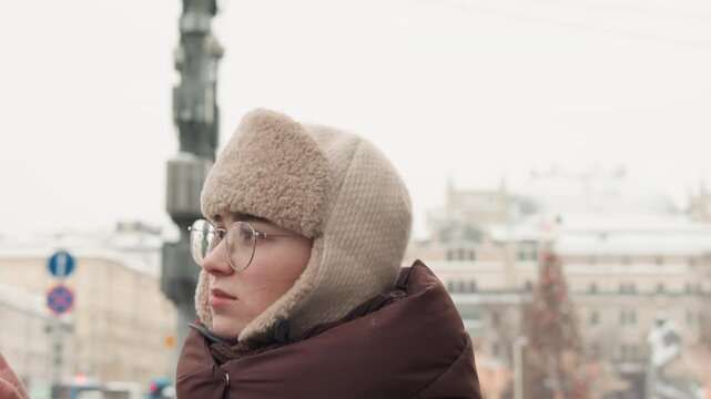 Young adult wearing knit trapper hat with shearling earflaps and puffy quilted jacket turns head, looking off frame with alert expression and visible breath in snowy urban street light