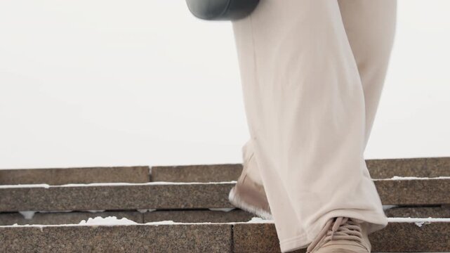 Low angle view of lower body descending outdoor stone steps, showing beige trousers and brown boots with snow on soles, raised heel, textured tread, and careful winter movement on rough surface