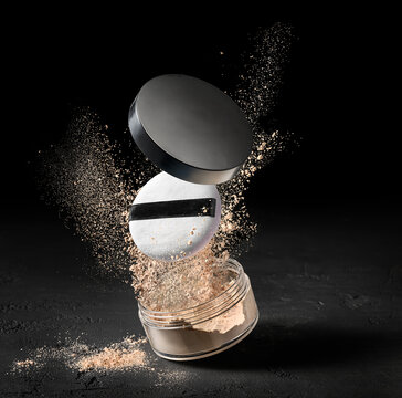Loose powder compact with open lid and makeup puff floating above sparkling beige particles on dark studio background