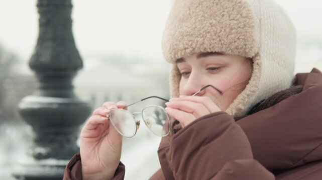 Young woman in fur lined trapper hat and padded winter jacket adjusts glasses and hat on snowy urban walkway, with calm side glance, soft smile, blurred city background and cool seasonal light