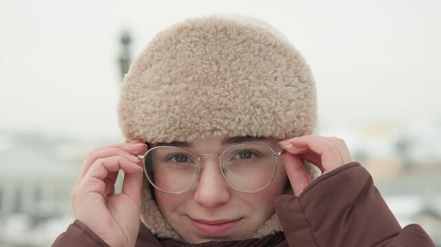 Frontal close portrait of young woman wearing beige fuzzy trapper hat and brown insulated coat as she lifts clear framed glasses, smiles gently, and stands in cold urban winter air