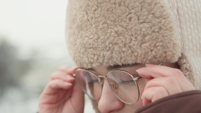 Close side-front view of young woman in shearling hat and hooded coat lifting eyeglasses into place with both hands while standing outdoors in chilly daylight, visible breath, flushed cheeks, knit