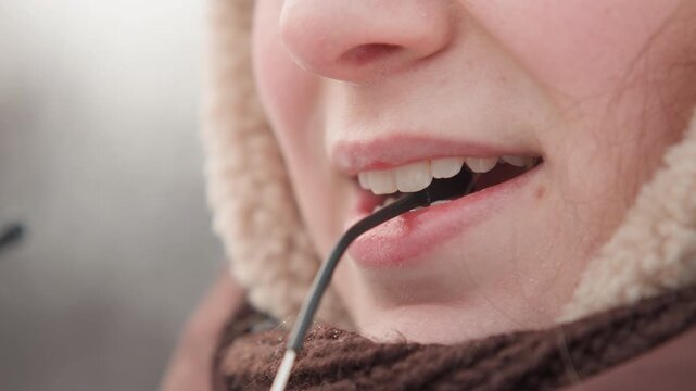 Tight frontal close-up of lower face wearing fur-lined hood and textured knitted scarf, holding eyeglass temple between teeth with lips slightly parted, subtle smile and relaxed winter mood outdoors