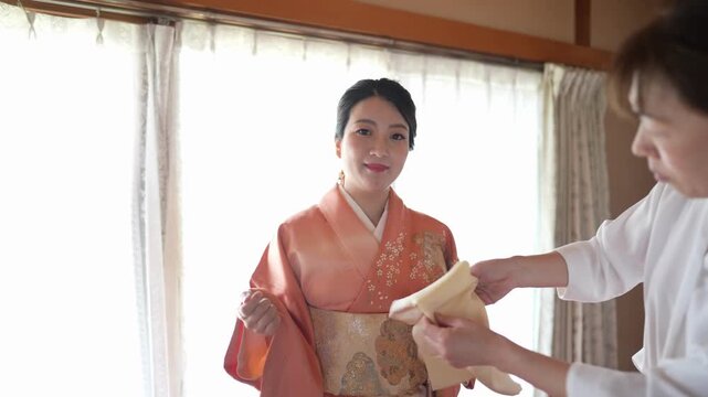 A woman in an orange kimono faces forward while an assistant holds a pale accessory beside her obi. Japan, April 19, 2025, spring.