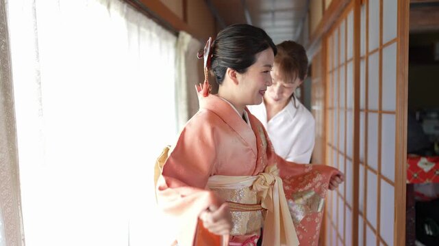 A smiling woman in an orange kimono stands beside sliding doors while an assistant works behind her. Japan, April 19, 2025, spring.