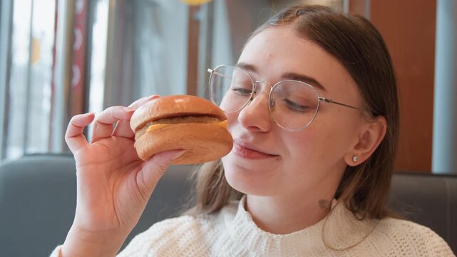 Young woman in cozy diner booth lifts burger toward face, inhales aroma, then moves sandwich toward mouth with slight smile, soft window light, relaxed posture, and warm casual meal atmosphere