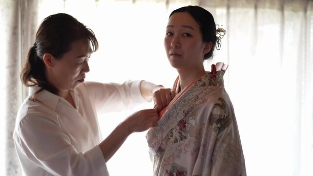 An assistant adjusts the collar of a woman in a patterned kimono as the woman looks toward the camera. Japan, April 19, 2025, spring.