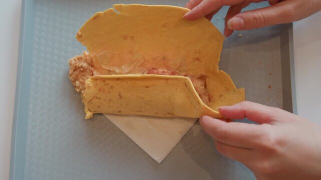 Top-down kitchen scene showing folded omelette on textured tray and napkin as hands lift and open it to expose shredded filling with small vegetable pieces, soft light and clean presentation