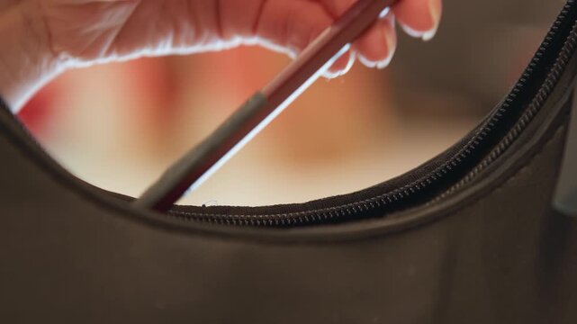 Close-up motion shows hand placing makeup brush into open zippered pocket of dark bag or garment, with gentle downward movement, curled fingers, soft diffused light, warm bokeh background