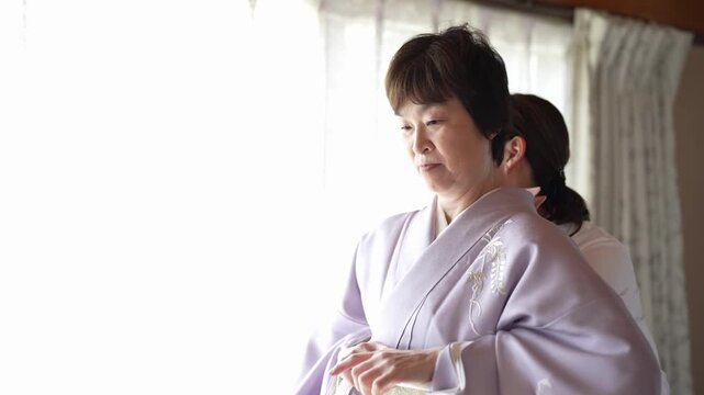 A woman in a lavender kimono holds her hands together while an assistant works behind her. Japan, April 19, 2025, spring.