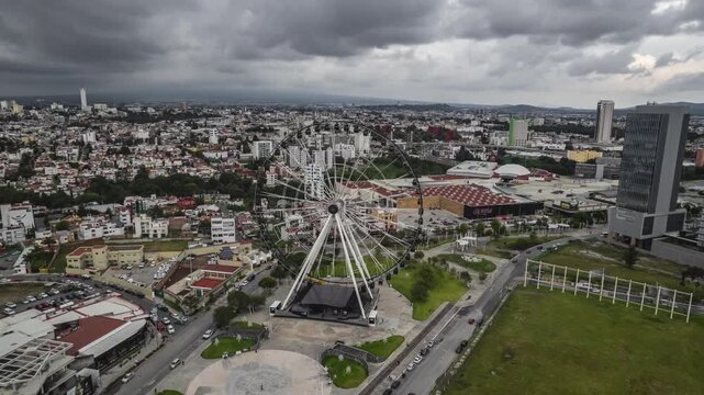Aerial drone footage of la estrella de puebla ferris wheel and cityscape under dramatic cloudy sky.