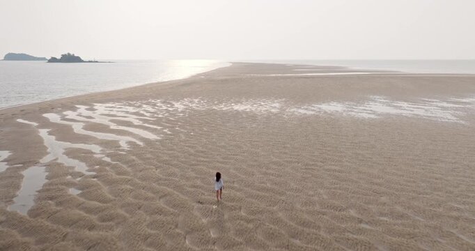 Woman exploring the vast sandy beach at low tide, showcasing unique ripple patterns and small pools of water with distant islands and a bright horizon in Ko Lanta, Thailand. Aerial view drone flight