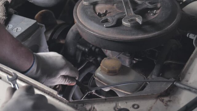 Mechanic repairs car engine with gloved hands in gritty workshop, focused and careful while tightening parts around air filter housing and coolant reservoir, automotive maintenance in close up.