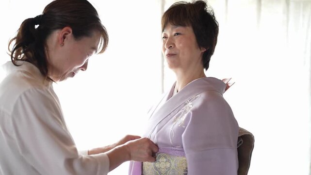 An assistant fastens the front cord on a patterned obi as a woman in a lavender kimono looks on. Japan, April 19, 2025, spring.
