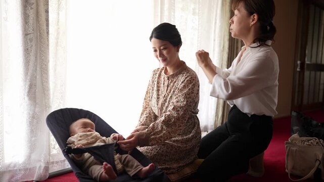 A woman extends her hands toward a baby in a bouncer while a hairstylist works on her hair. Japan, April 19, 2025, spring.