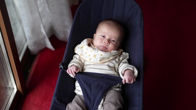 An alert baby in striped clothing and a star-patterned bib reclines in a navy bouncer by a window. Japan, April 19, 2025, spring.