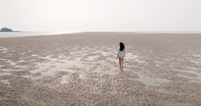 Woman in white t-shirt walking barefoot on vast sandbar at low tide, finding solitude and peaceful contemplation in the expansive, remote coastal landscape of Ko Lanta, Thailand. Tracking drone flight