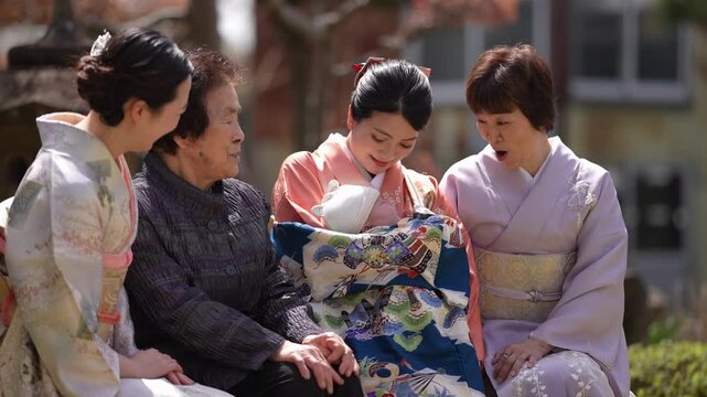 Four women sit close together in a garden and look toward a baby draped in a colorful patterned garment. Japan, April 19, 2025, spring.