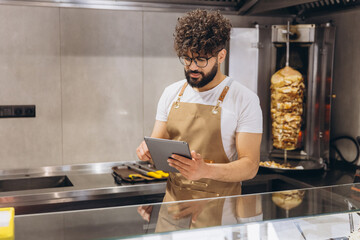 Chef in a restaurant kitchen using a tablet to manage orders and operations, with a rotating shawarma skewer in the background © anatoliycherkas