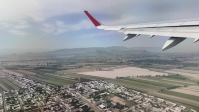 An airplane is taking off with a view from the porthole. The landscape of Osh city in Kyrgyzstan is visible below. Fields and buildings appear as the flight ascends into the sky.